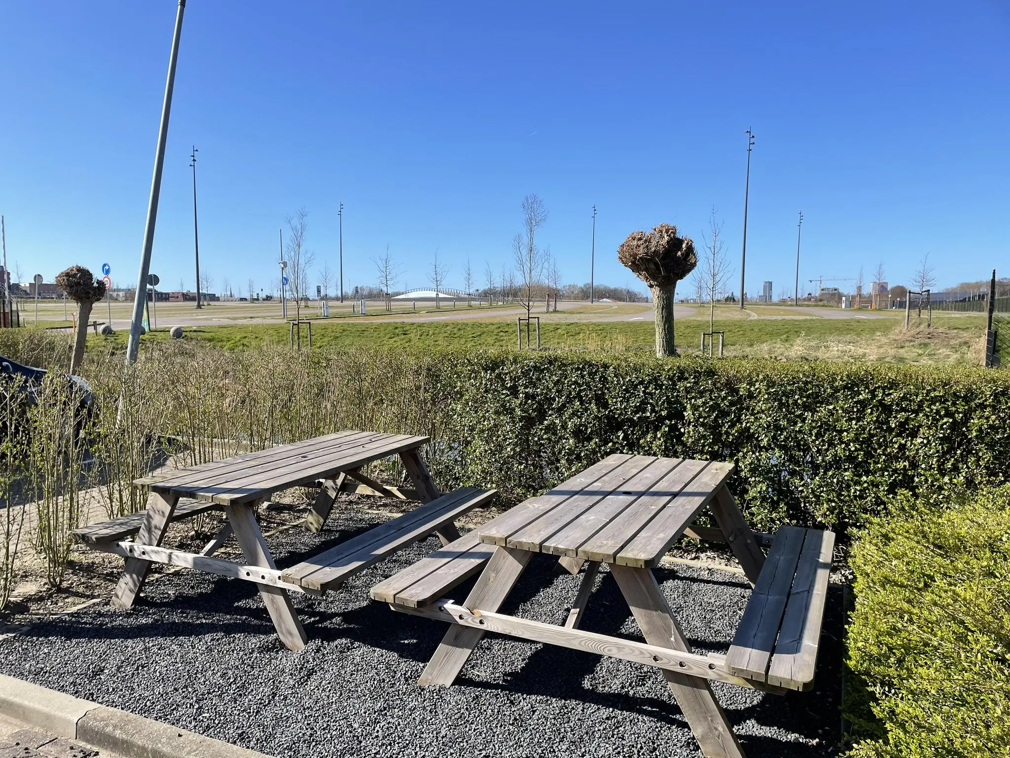 Twee picknicktafels staan naast een heg aan de Laan van ’s-Gravenmade met uitzicht op een open grasveld en blauwe lucht.